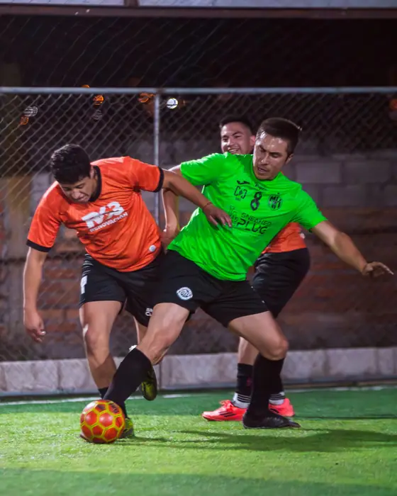 Choque por el balón entre un jugador de naranja y uno de verde